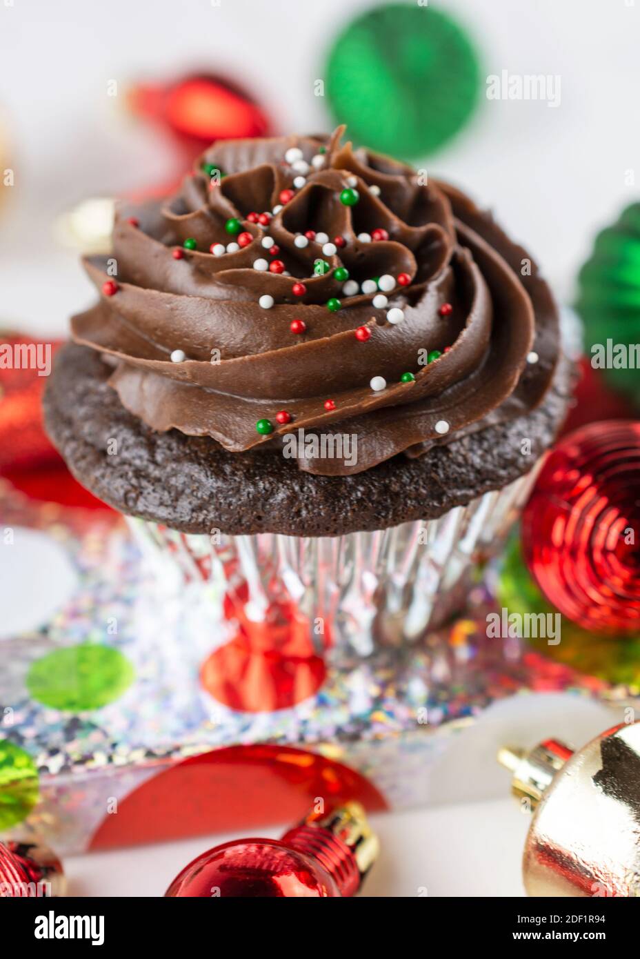 A Chocolate Christmas cupcake sits on a wrapped present surrounded by green, gold, and red ornaments.  White background. Stock Photo