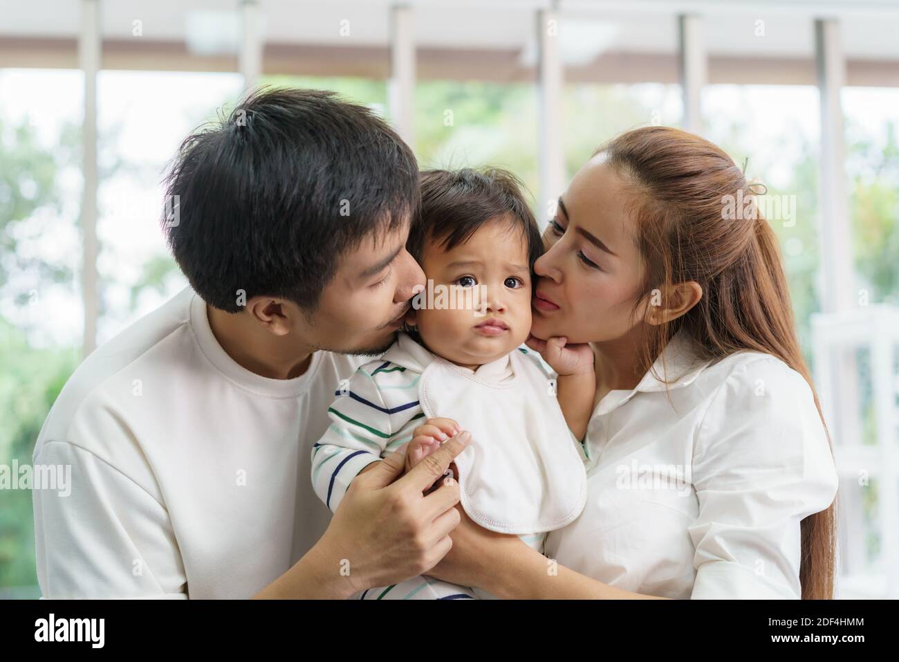 Asian family happy kiss little baby boy in living room at home, young parent and children enjoy love hug standing together. Stock Photo