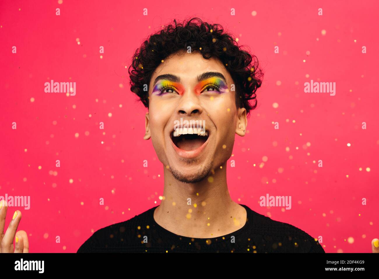 Androgynous male with rainbow eye makeup throwing up the glitter and laughing. Excited gay man with glitters flying against red background. Stock Photo
