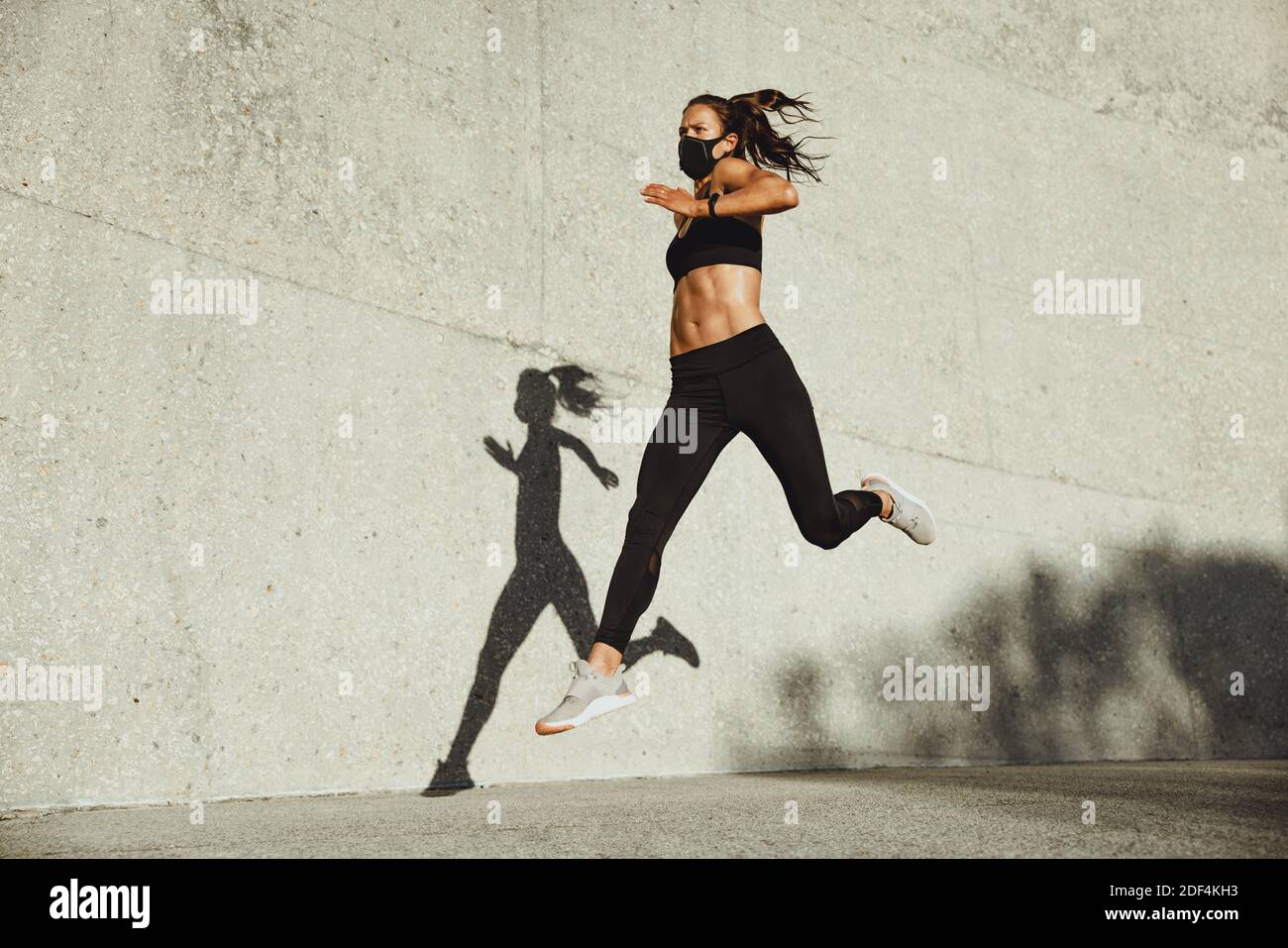 Athletic woman with face mask running outdoors. Female runner in sports clothing and protective face mask sprinting outdoors. Stock Photo
