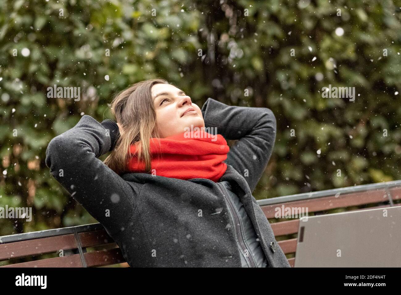 A young woman, with her hands behind her head, feels peace, rests after work, sits on a wooden bench in the park a laptop on her knees. Break during w Stock Photo