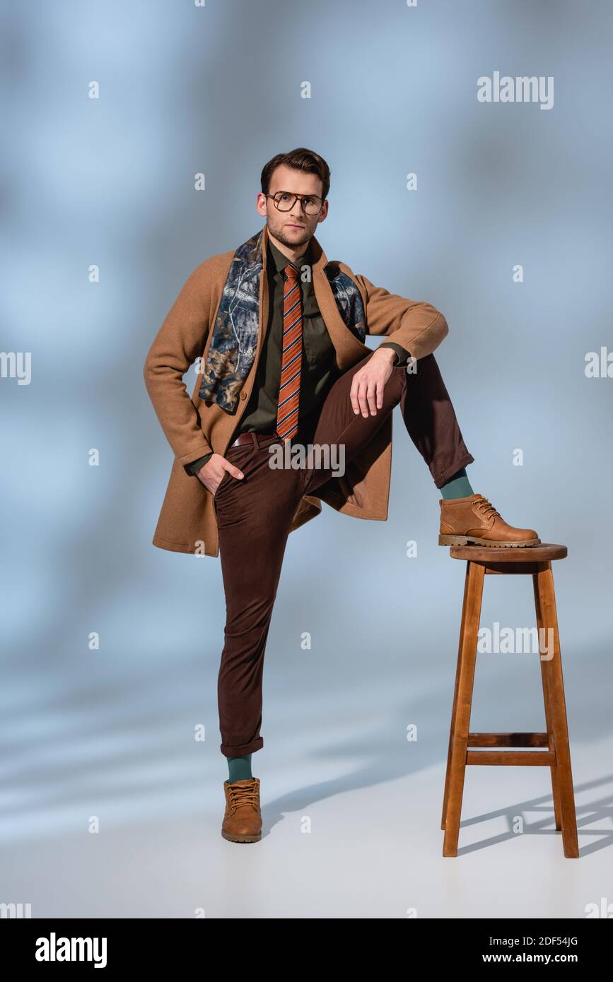 full length of stylish man in winter outfit and glasses posing while leaning on wooden stool on grey Stock Photo