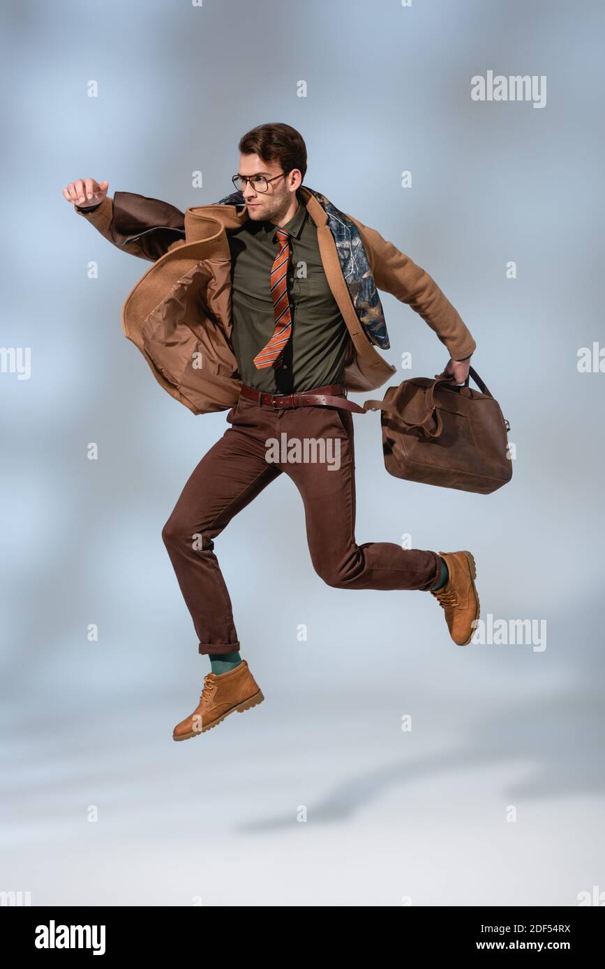 stylish man in winter coat holding leather briefcase while jumping on grey Stock Photo