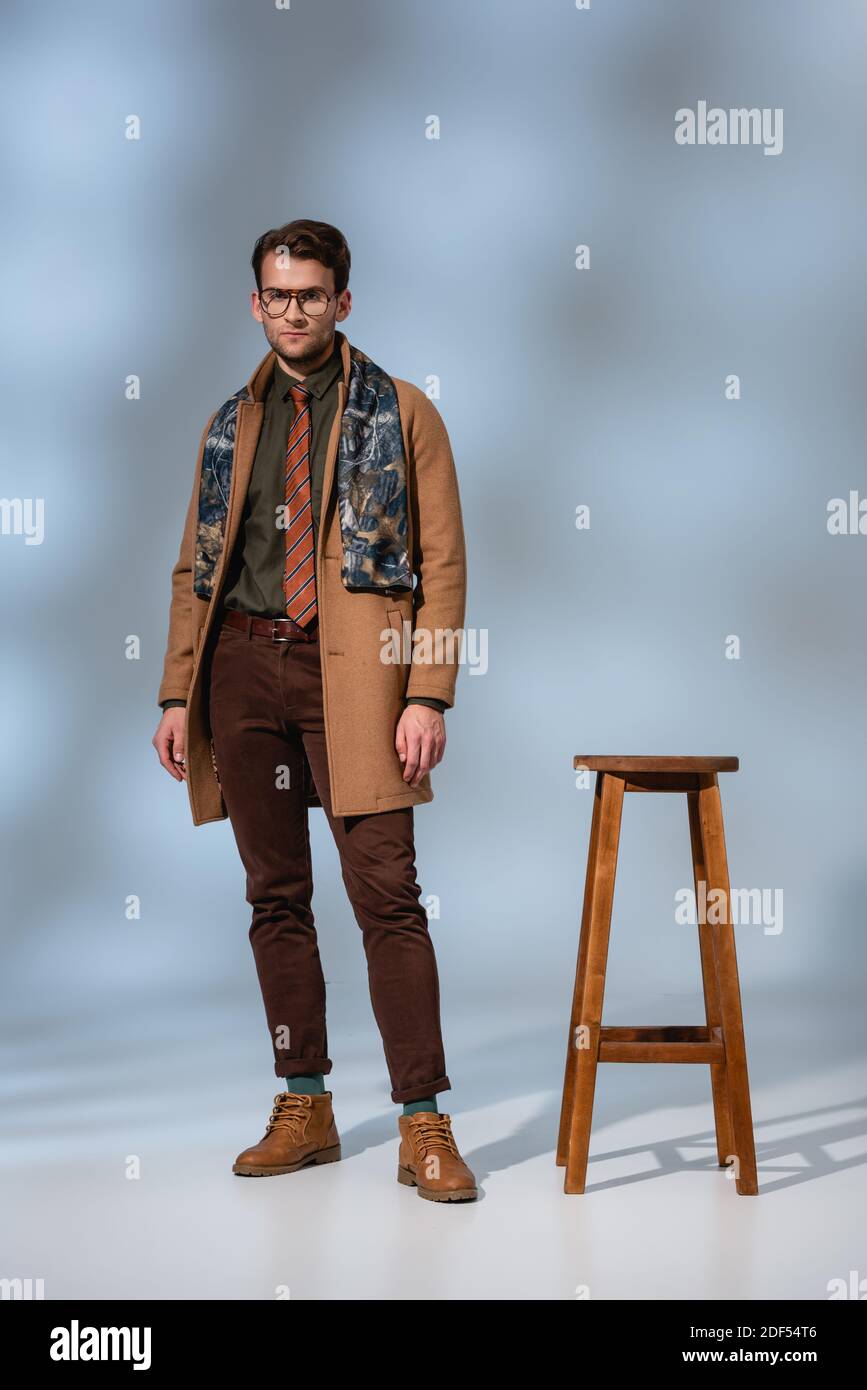 full length of stylish man in winter outfit and glasses standing near wooden stool on grey Stock Photo