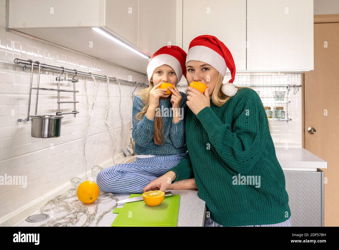 Cute little girl and her beautiful mother in Santa hats are looking at camera and smiling with oranges slices. Stock Photo