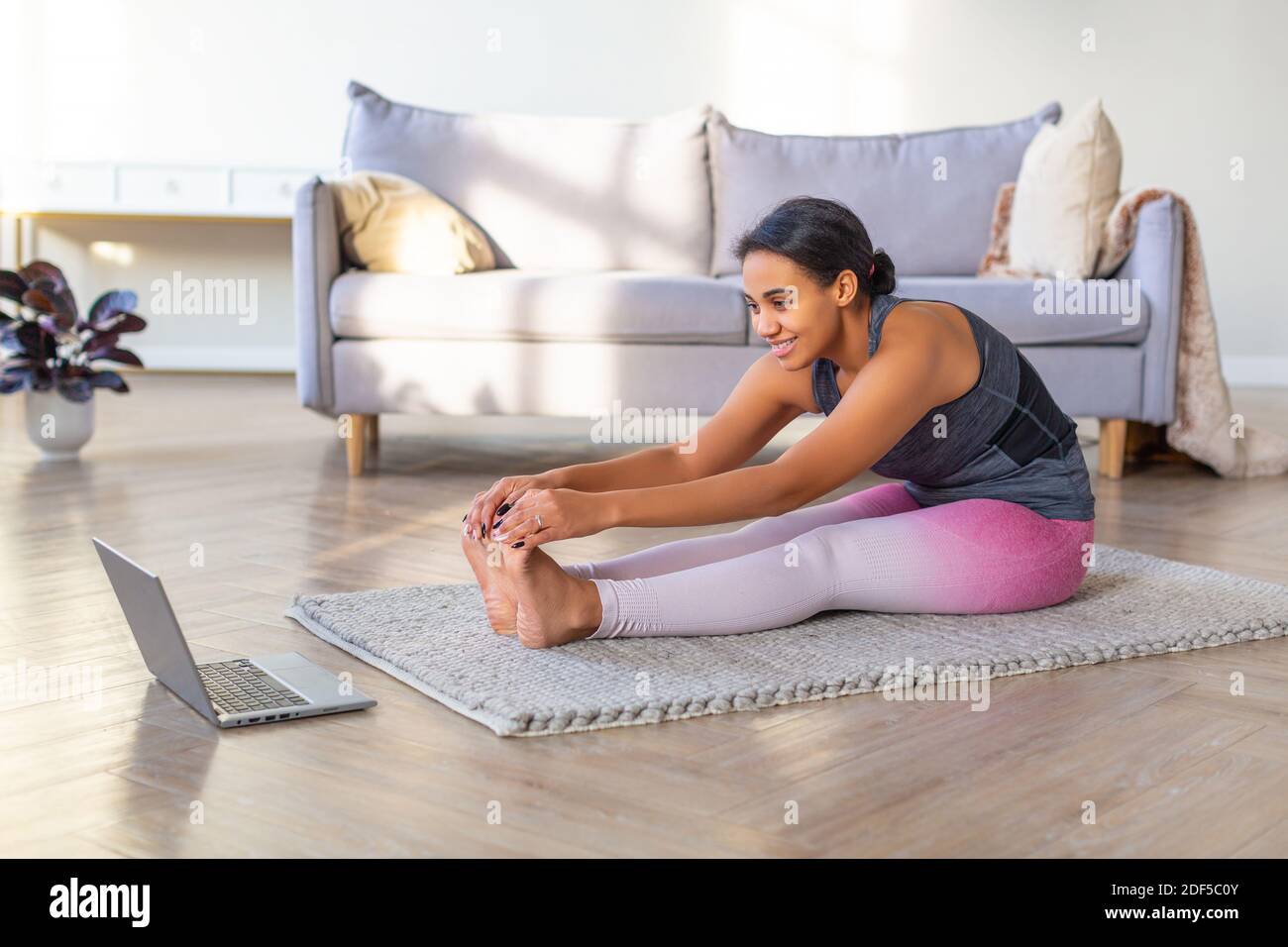 Smiling African-american woman doing stretching at home. She sits on a training mat in front of a laptop monitor. Stock Photo