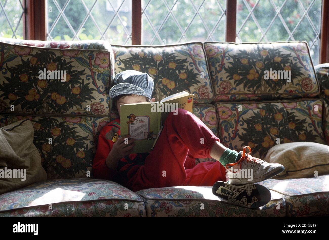 Young boy aged six wearing a Osh Kosh hat and  reading a Disney Peter Pan & Wendy book whilst seated on a settee as November sunlight comes through the leaded windows of his home in Englland. Stock Photo