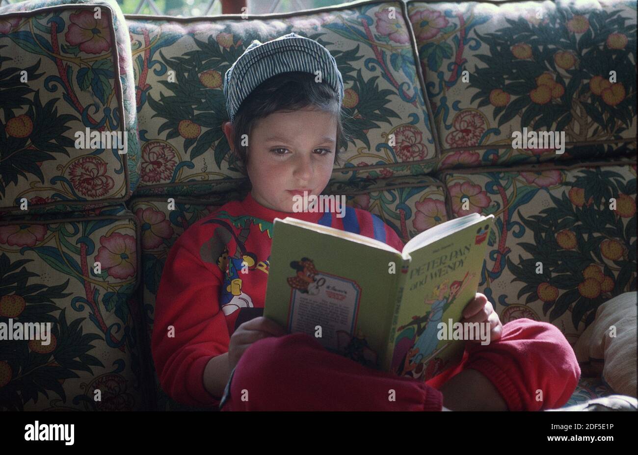 Young boy aged six wearing a Osh Kosh hat and  reading a Disney Peter Pan & Wendy book whilst seated on a settee as November sunlight comes through the leaded windows of his home in Englland. Stock Photo