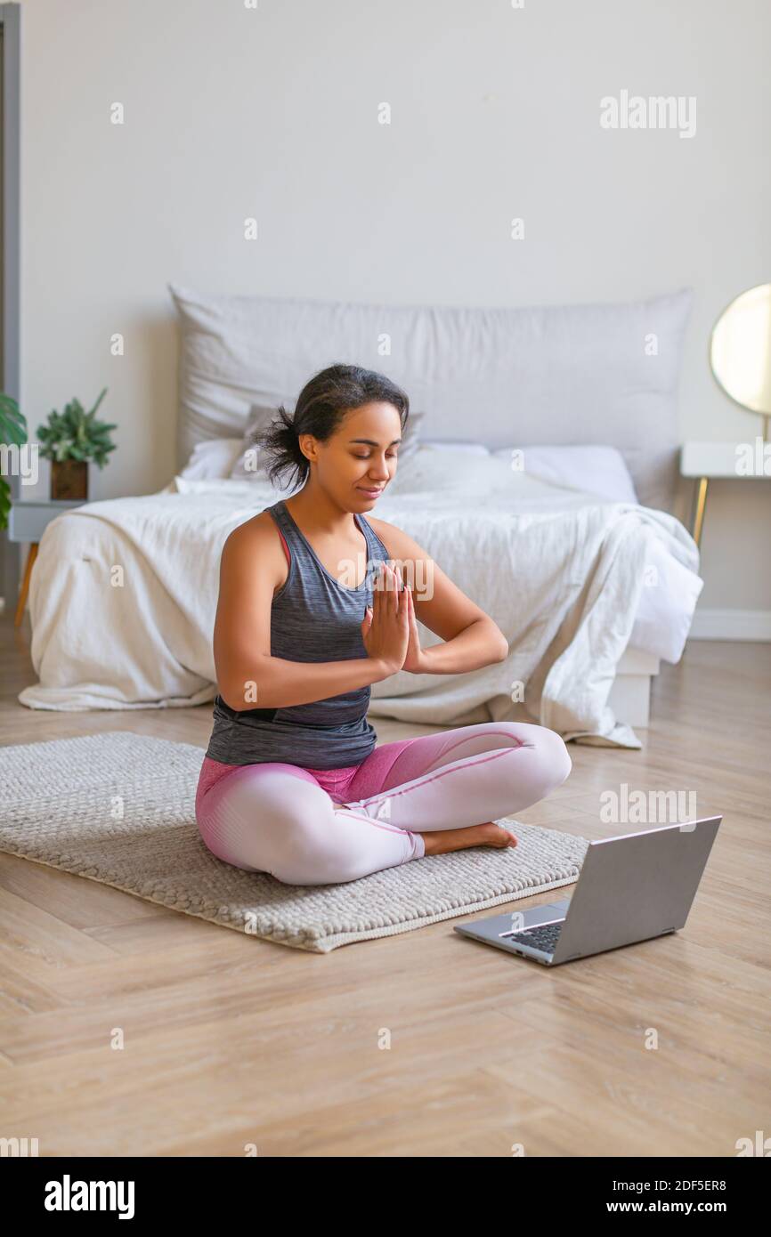 Vertical photo of an African-american woman meditates at home in front of a laptop monitor with online tutorials. Stock Photo