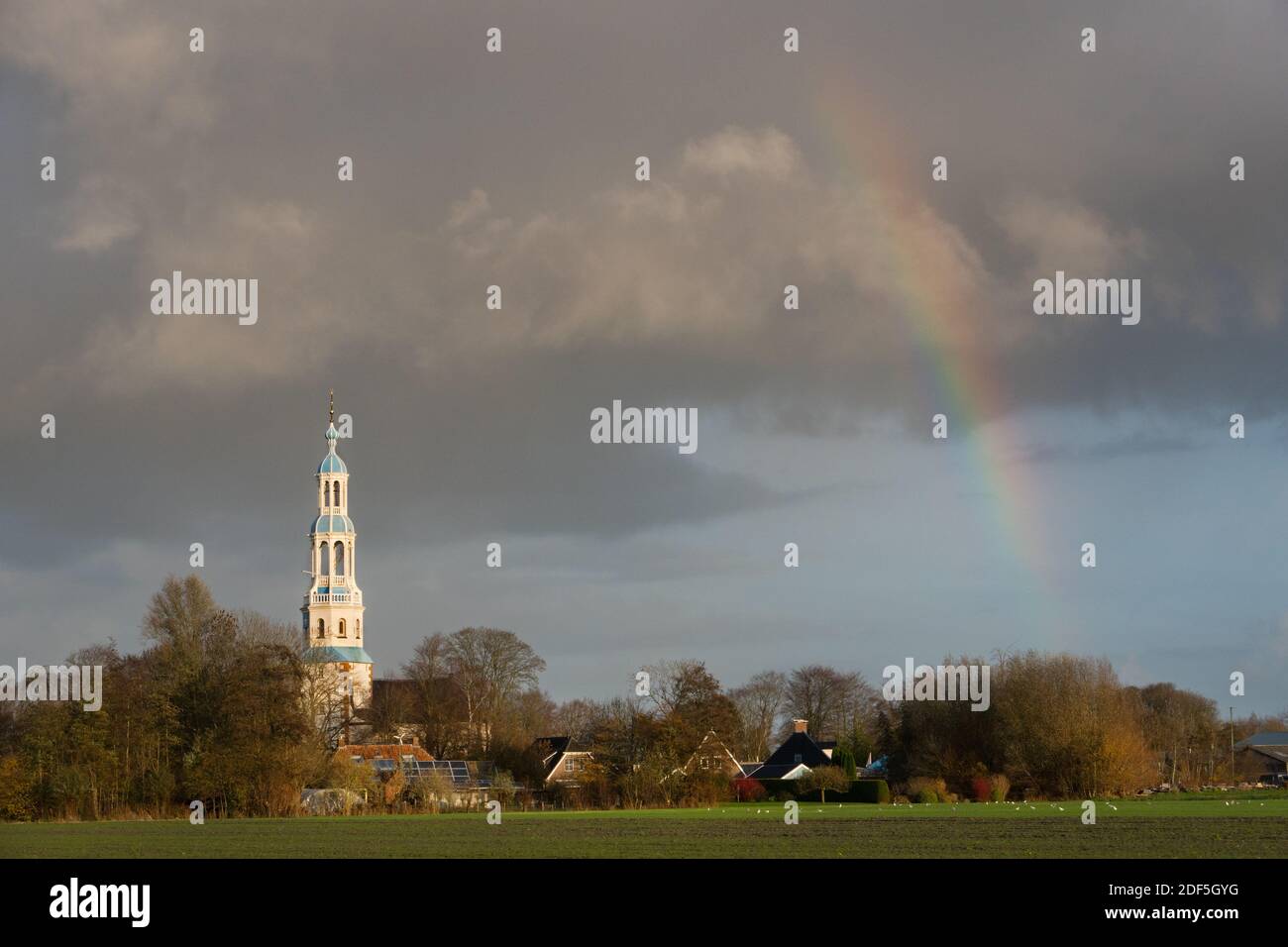 Small rural village with white church tower under dark clouds with rainbow Stock Photo