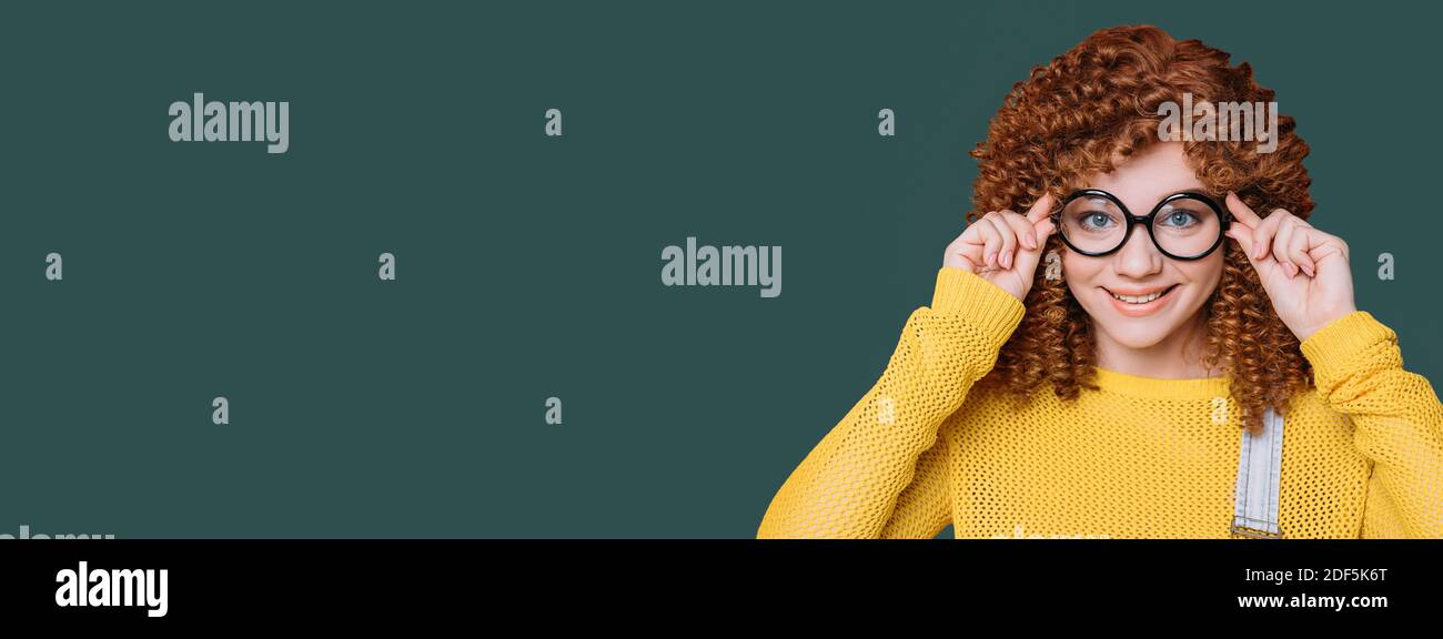 Cheerful red-haired curly woman holding trendy glasses near her face, isolated over tidewater green background, empty for text Stock Photo