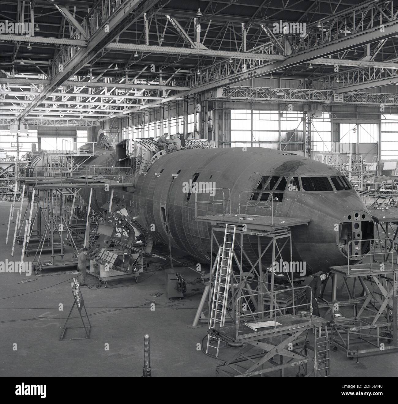 1950s, historical, inside a large aviation hangar, a commerical aircraft being constructed, England, UK. Picture shows the nose or cockpit, with technicans working on the foor and on top of the aeroplane. Stock Photo