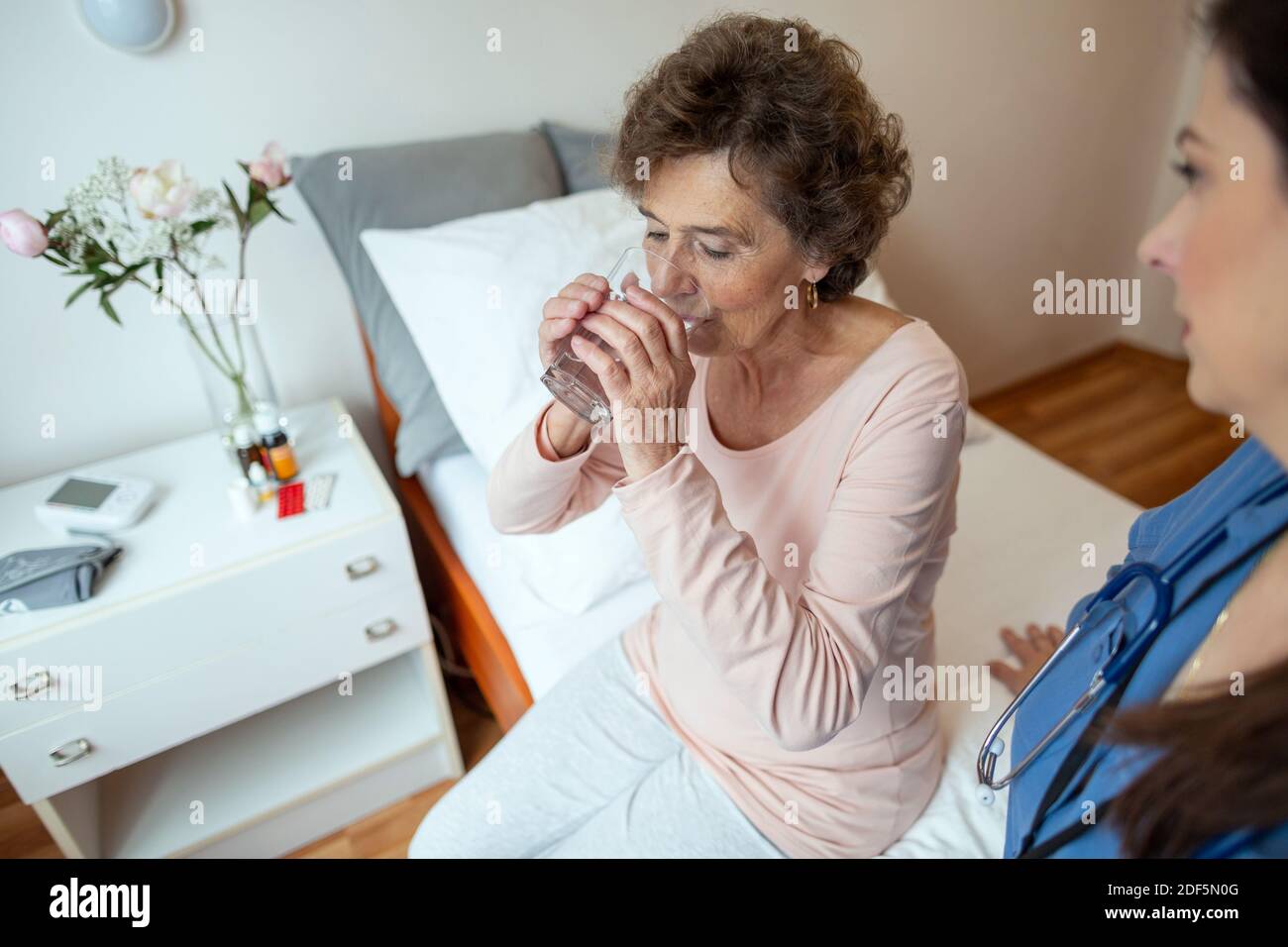 Senior Woman Patient Drinking Water. Elderly Female Patient Sitting on Hospital Bed with Nurse Watching Her. Stock Photo