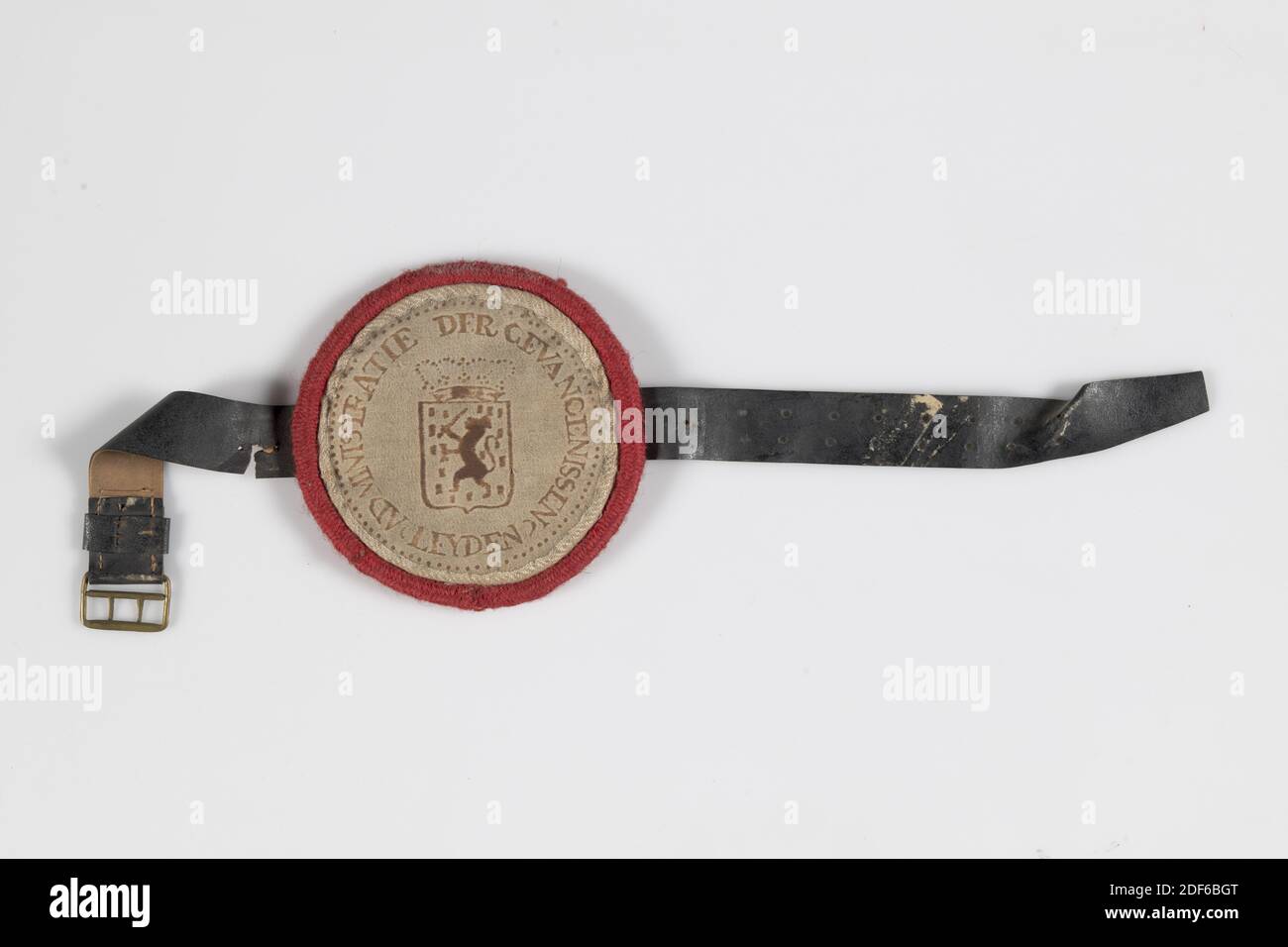 bracelet insignia, Anonymous, first half of the 19th century, cotton, linen, leather, brass, wool, sewn, General: 12 x 45 x 0.8cm 120 x 450 x 8mm, weapon sign, Bracelet intended as a way of passing through a fire in prison, consisting of a white cotton insignia with the inscription ADMINISTRATION OF THE PRISONS LEYDEN. Within the inscription is the national coat of arms of the Netherlands, consisting of a crowned shield with a crowned lion climbing with a sword. A white linen edge is sewn around the insige with a red wool edge around it. The back of the insignia is plain. A black leather strap Stock Photo