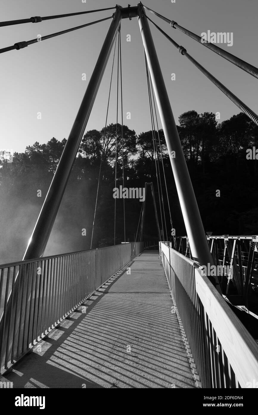 A pedestrian suspension bridge catching the last rays of the sun. Spray from a waterfall drifts into the photo from the left. Black and white Stock Photo