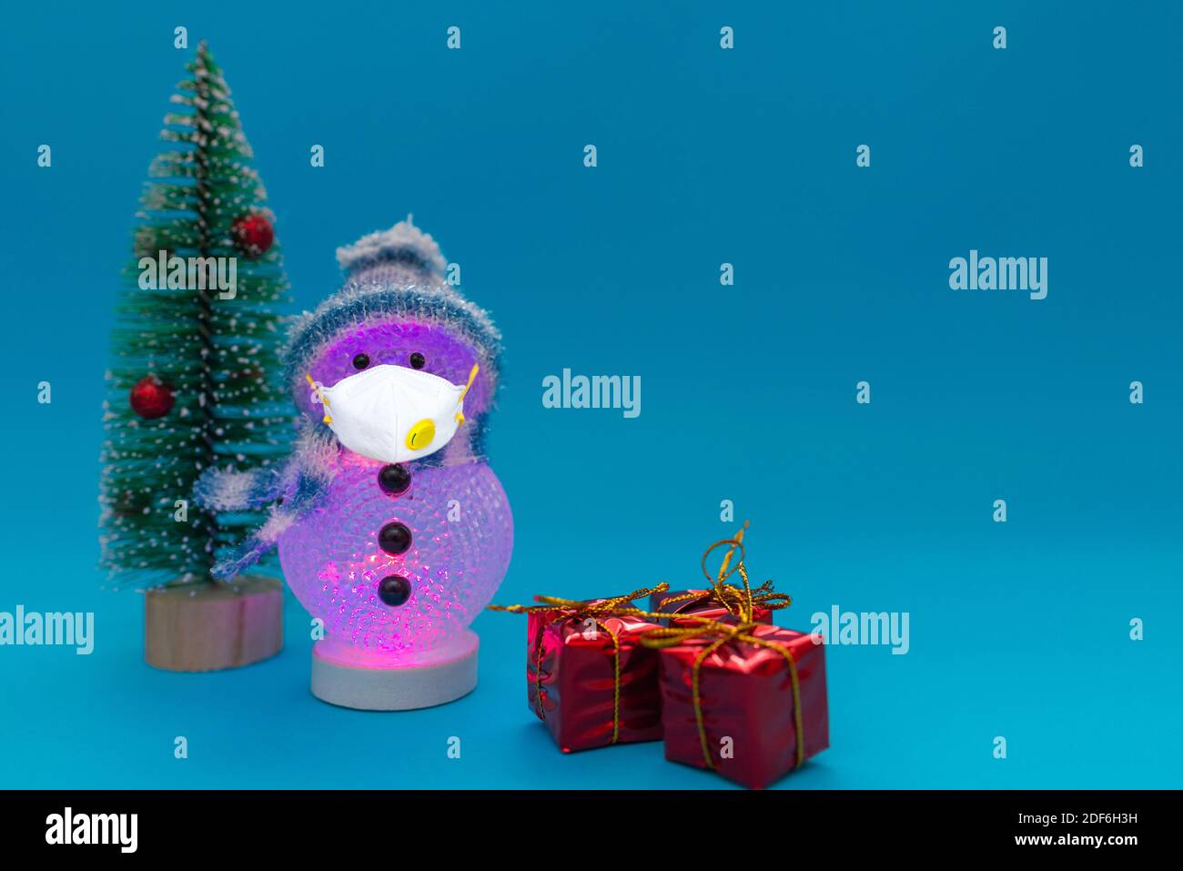 Glowing snowman at evening wears face mask, presents and Christmas tree with copy space.Christmas and new year celebration.Studio shot. Stock Photo