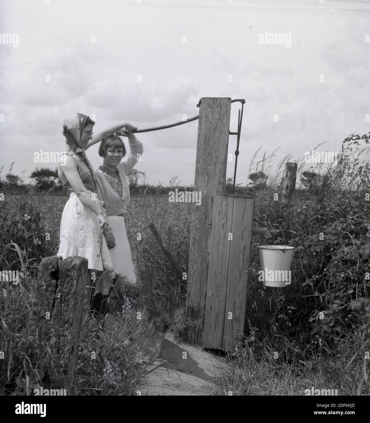 1940s, historical, on a narrow path beside a field, a mother and daughter pushing the wooden handle of a water well to pump water into a metal bucket, England, UK. Growing up in the countryside or a farm in this era meant that a youngster would be expected to help and support her parents on all kind of different jobs. Stock Photo