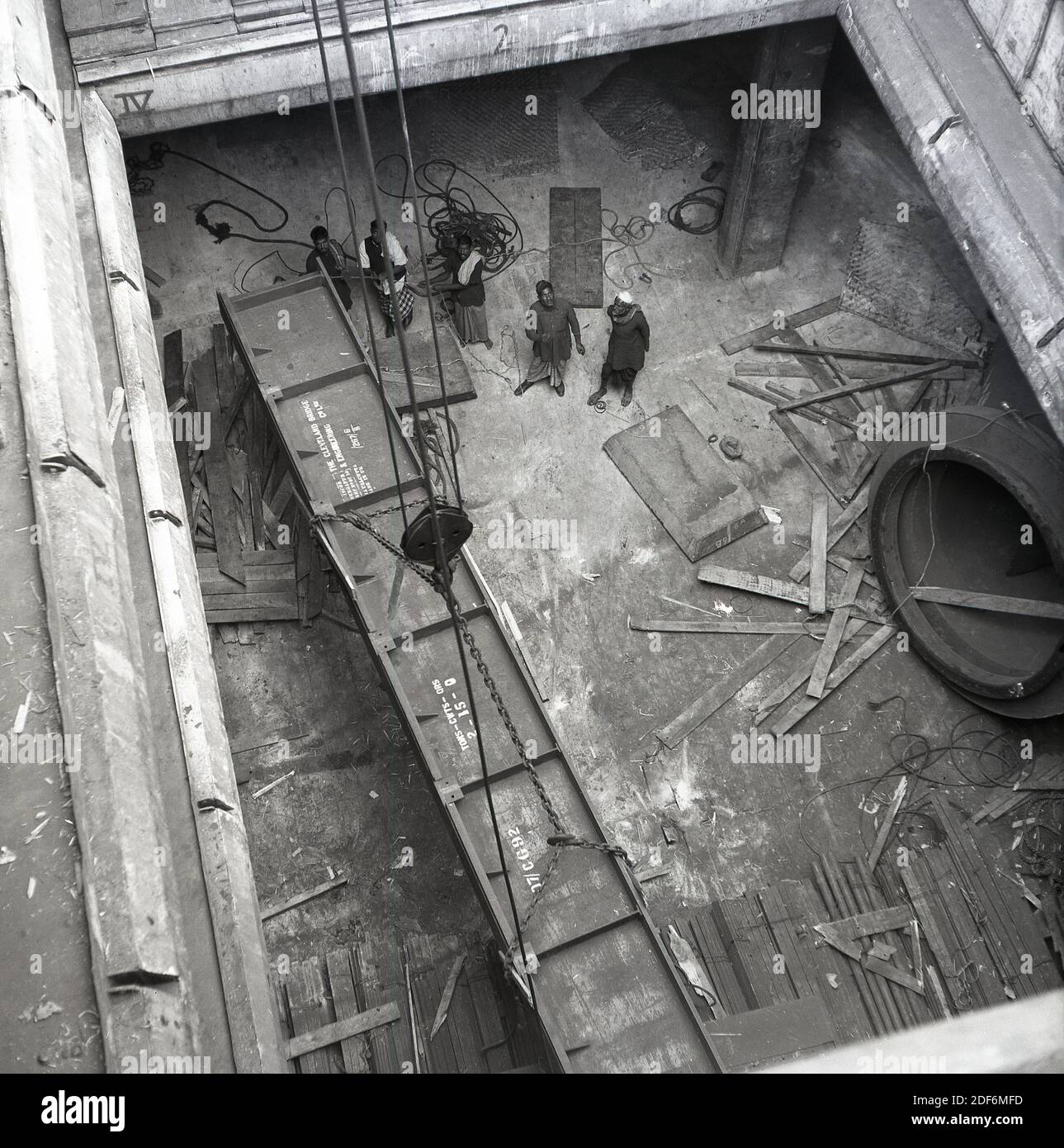 1950s, historical, India, Bombay, overhead view looking down at the cargo hold of a container ship and indian workers watching as a large block of heavy metal is lifted up. The metal freight is two tons in weight and not a hard hat in sight. Some of the port workers are even in barefeet or wearing light sandals. Stock Photo