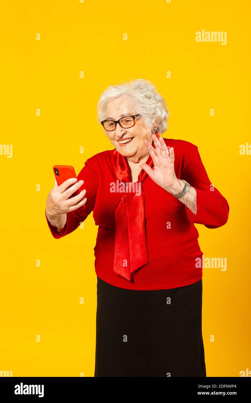 Studio portrait of a senior woman wearing a red shirt against a yellow background and having a video call with a red mobile phone Stock Photo