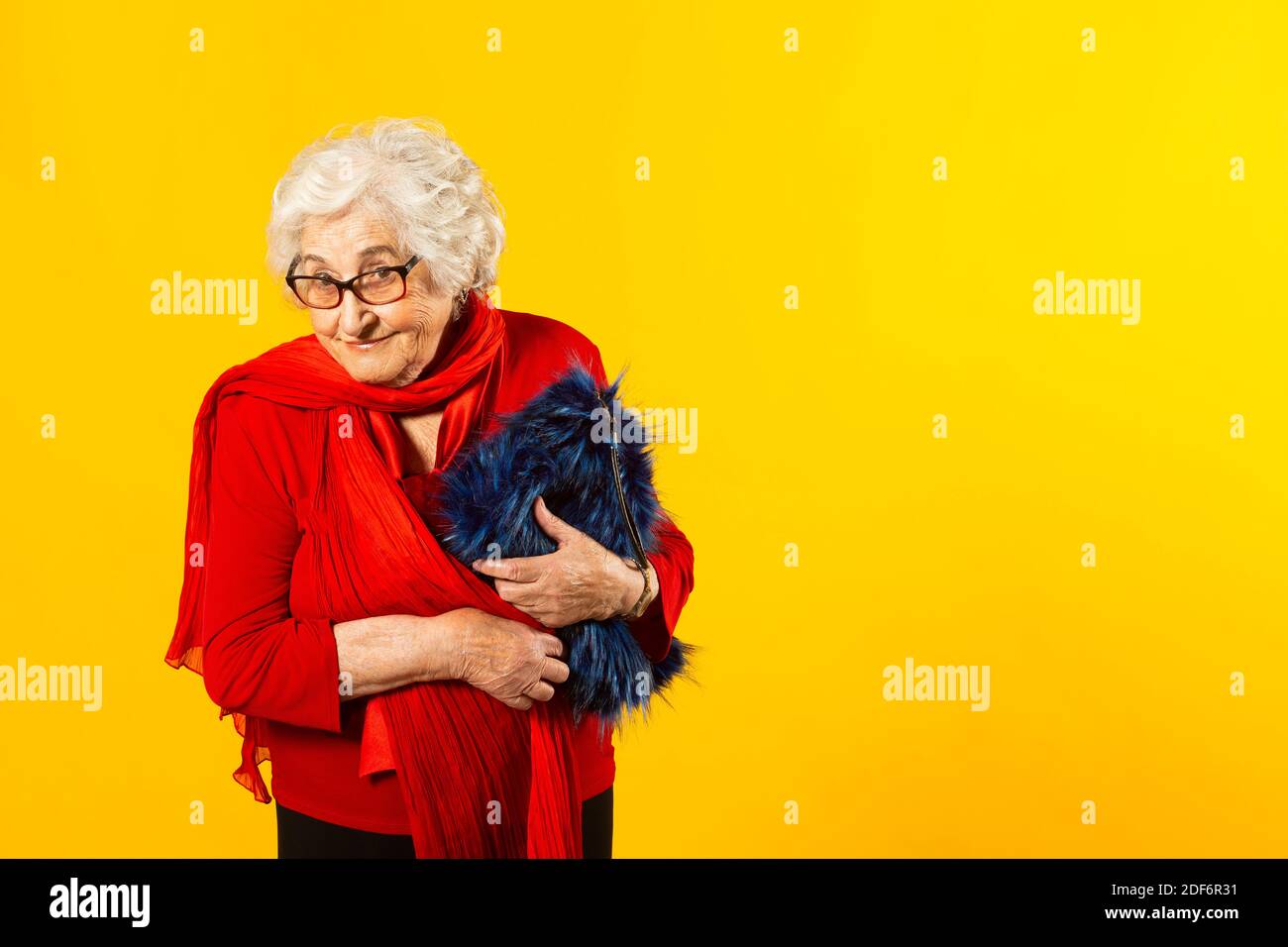 Studio portrait of a senior woman wearing a red shirt and a blue hairy bag, against a yellow background Stock Photo