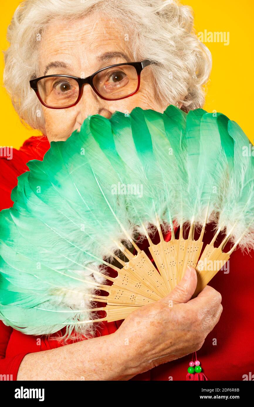 Studio portrait of a senior woman wearing a red shirt and a green feather hand fan, against a yellow background Stock Photo