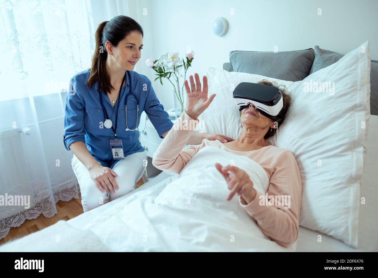 Female Doctor Checking on Elderly Patient Lying in Hospital Bed Doing Therapy Via VR Technology. Friendly Doctor Helping Senior Woman Receive Therapy. Stock Photo