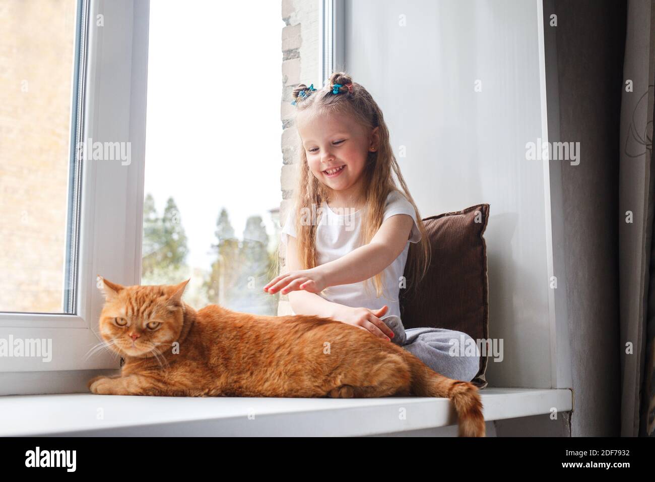 Little girl in pajamas relaxing on a window sill with red cat. Happy weekend with cat at home. Stock Photo