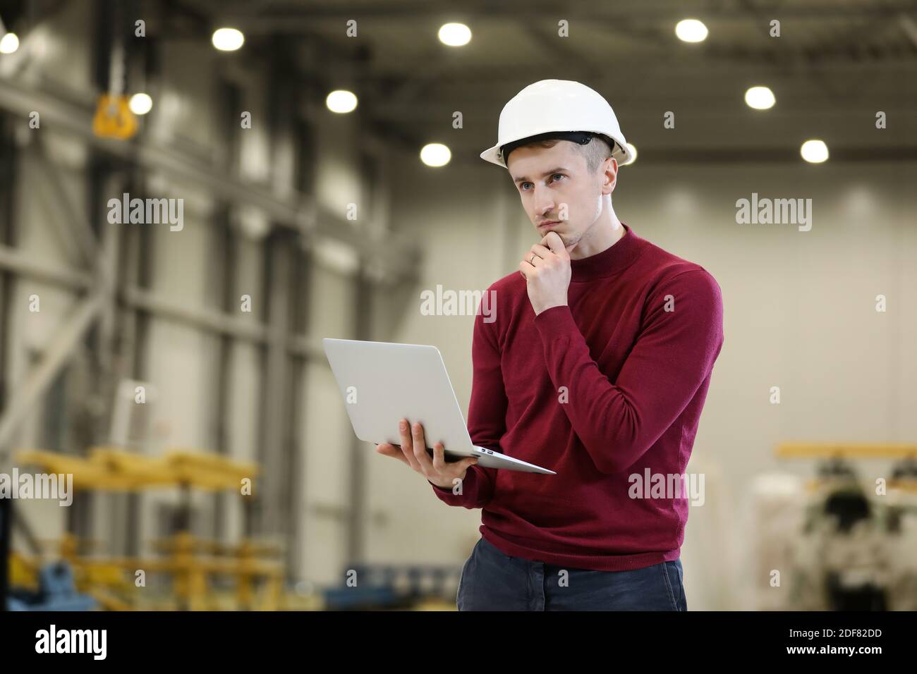 Portrait of a male factory manager in a white hard hat and red sweater holding laptop and mobile phone. Controlling the work process in the helicopter manufacturer. . High quality photo Stock Photo