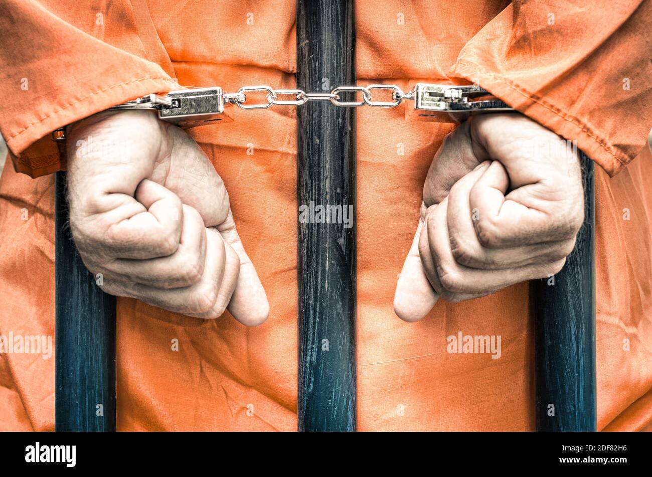 Handcuffed hands of a prisoner behind the bars of a prison with orange clothes - Crispy desaturated dramatic filtered look Stock Photo