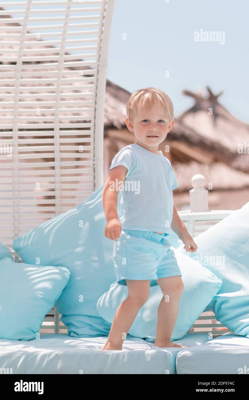 Kid boy blond on vacation in cafe by sea stands and look at camera. Vertical shot, lifestyle about vacation and travel with children. Stock Photo