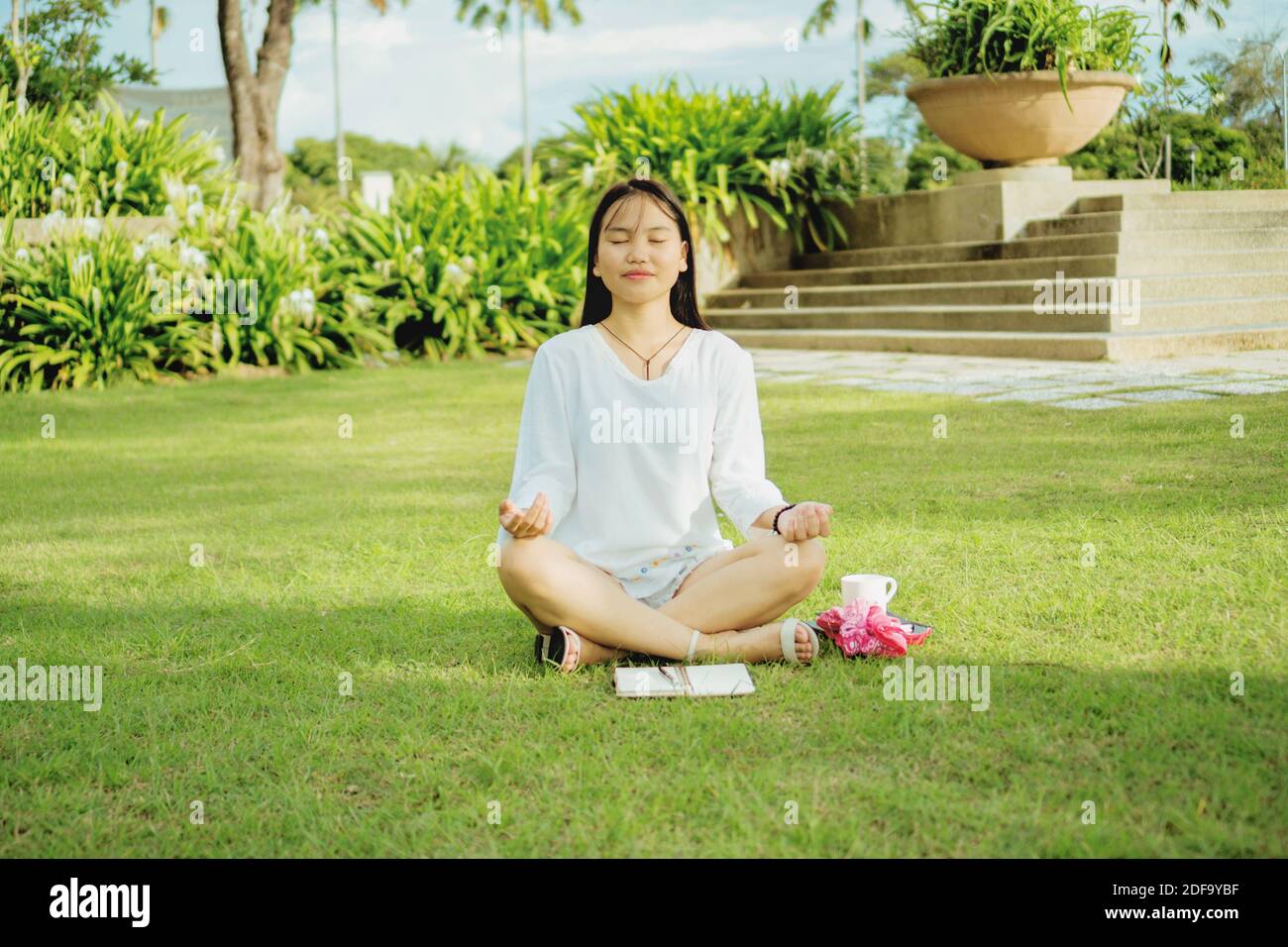 Young asian woman sitting with legs cross on the grass in a public park and meditating. Outdoor lifestyle photography. Stock Photo