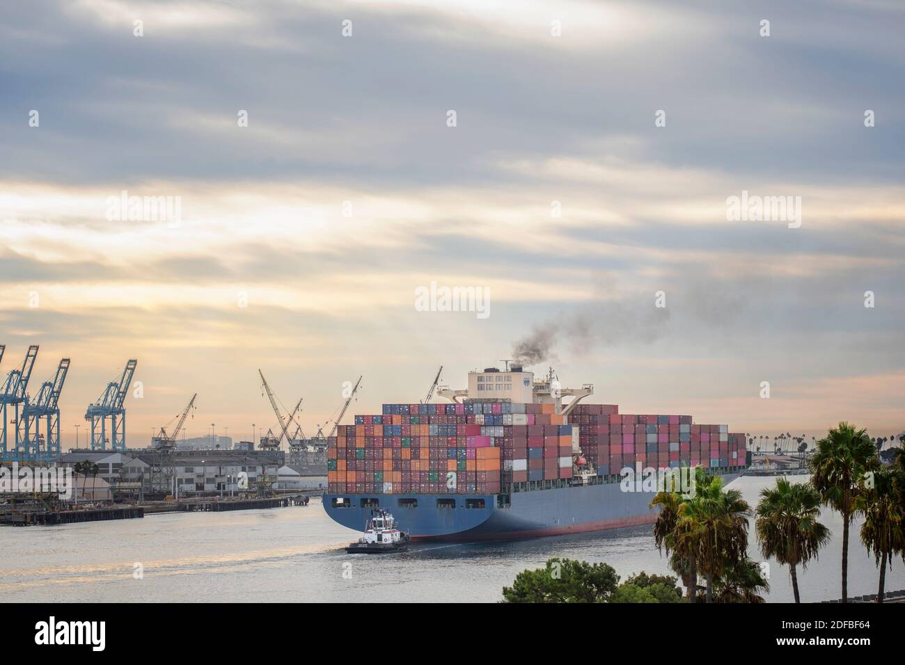 A huge freight container ship sailing into the ocean under dramatic sky emitting dark smoke of engine pollution in the air Stock Photo