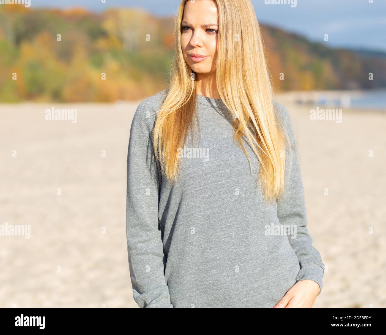 Blond woman in grey blouse is standing on beach and looking on side. She is posing outdoor for fashion mockup. Stock Photo