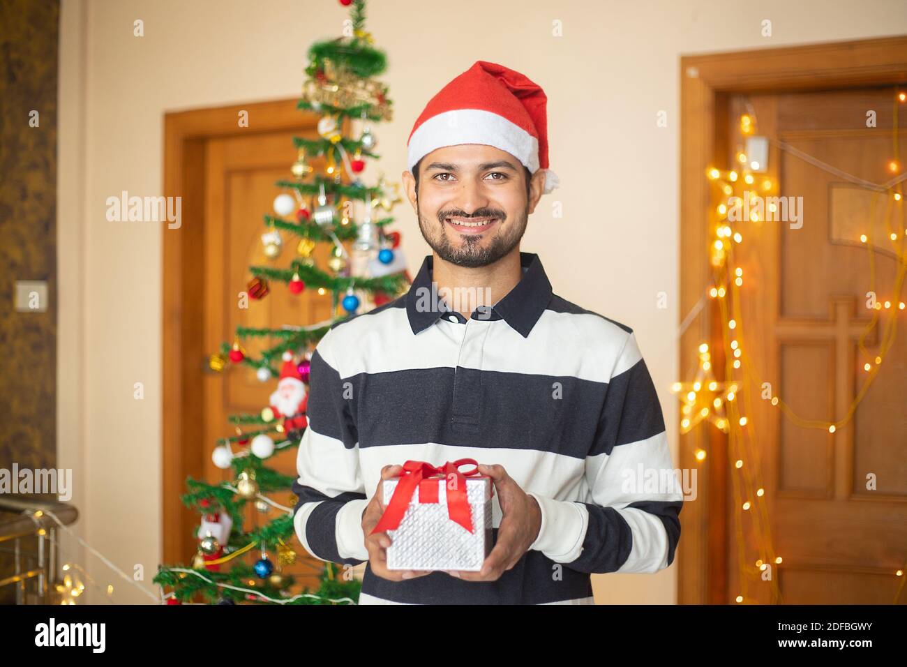 Cheerful Young Indian man holding gift box in hand celebrating christmas alone at home, new year, holiday, party, winter, december. quarantine due to Stock Photo