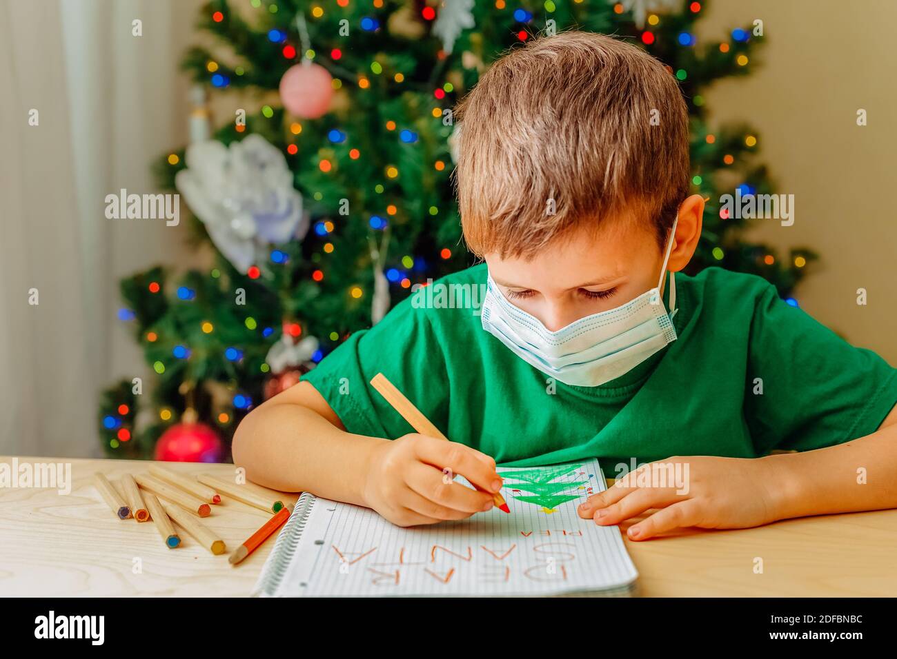 Smiling 7 years old boy wearing green t-short write letter to Santa sitting by desk, Christmas tree on background Stock Photo