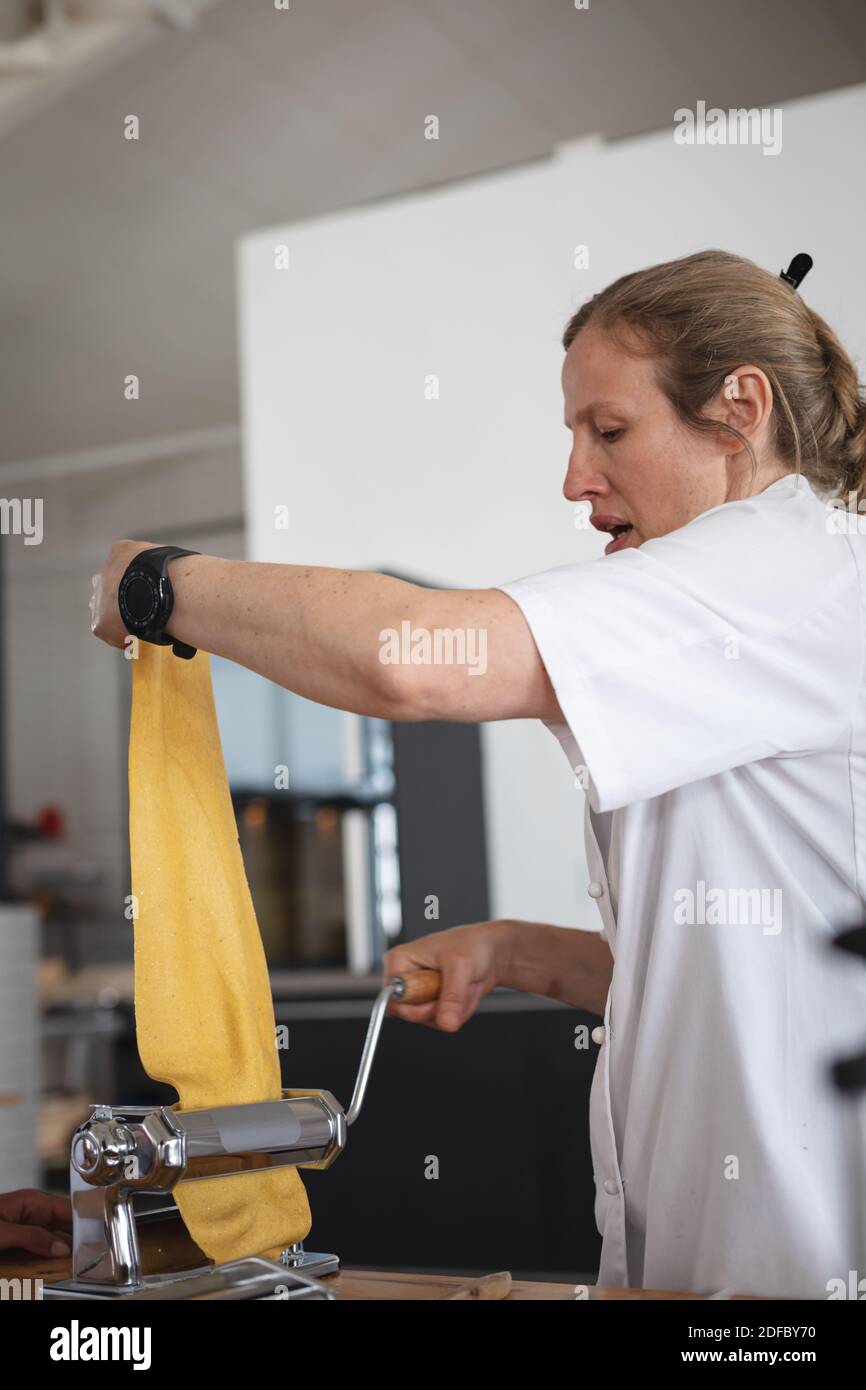 Senior caucasian female chef processing pasta sheet in machine at restaurant kitchen Stock Photo