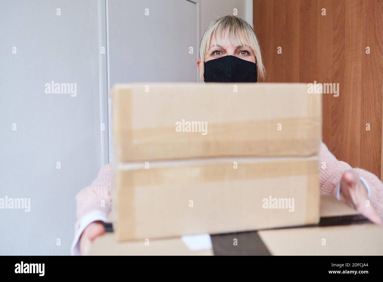 Woman with face mask holds packages in her hands in front of the door of her apartment Stock Photo