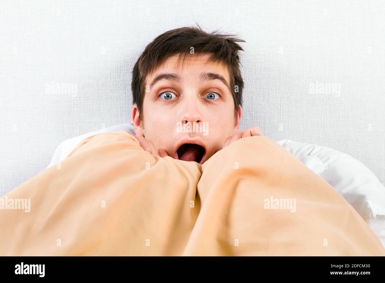 Surprised Young Man under Blanket in the Bed at the Home Stock Photo