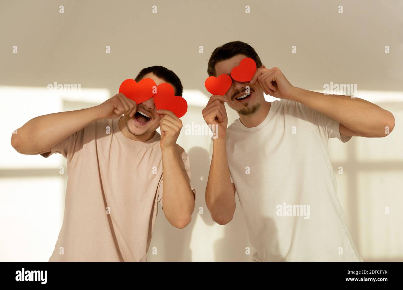 Happy couple of young gay men hide their eyes behind red paper valentine hearts in celebration at home against white wall background Stock Photo