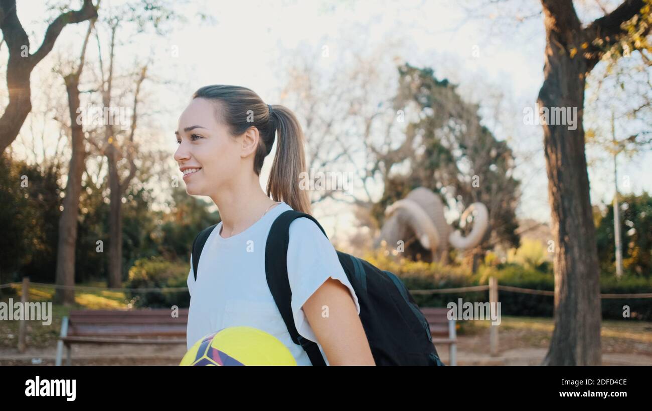 Beautiful girl with backpack and football walking through city park ...