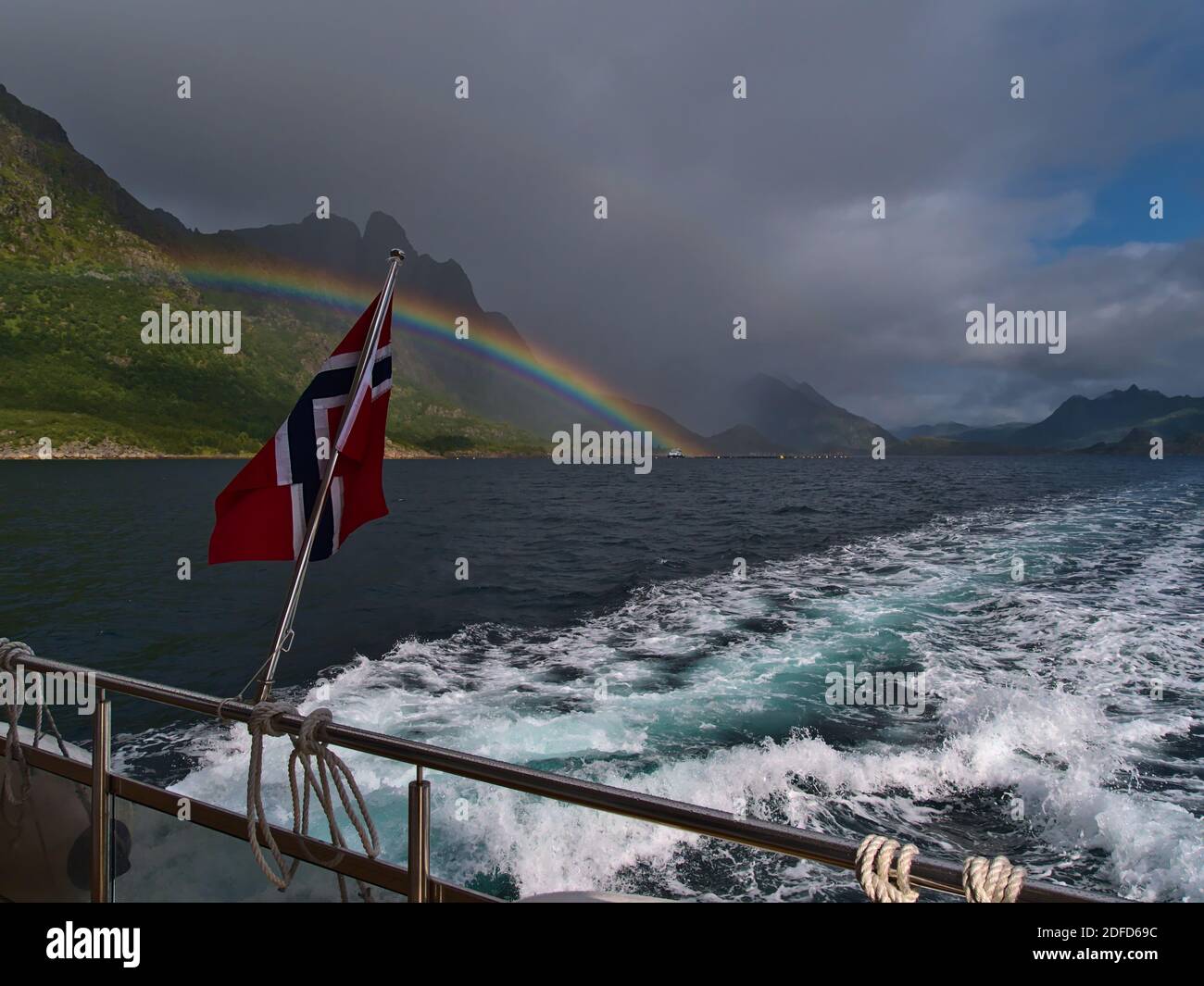 Norwegian national flag flying in the wind on the stern of tourist boat with majestic colorful rainbow above mountains on the rocky coast of Norway. Stock Photo