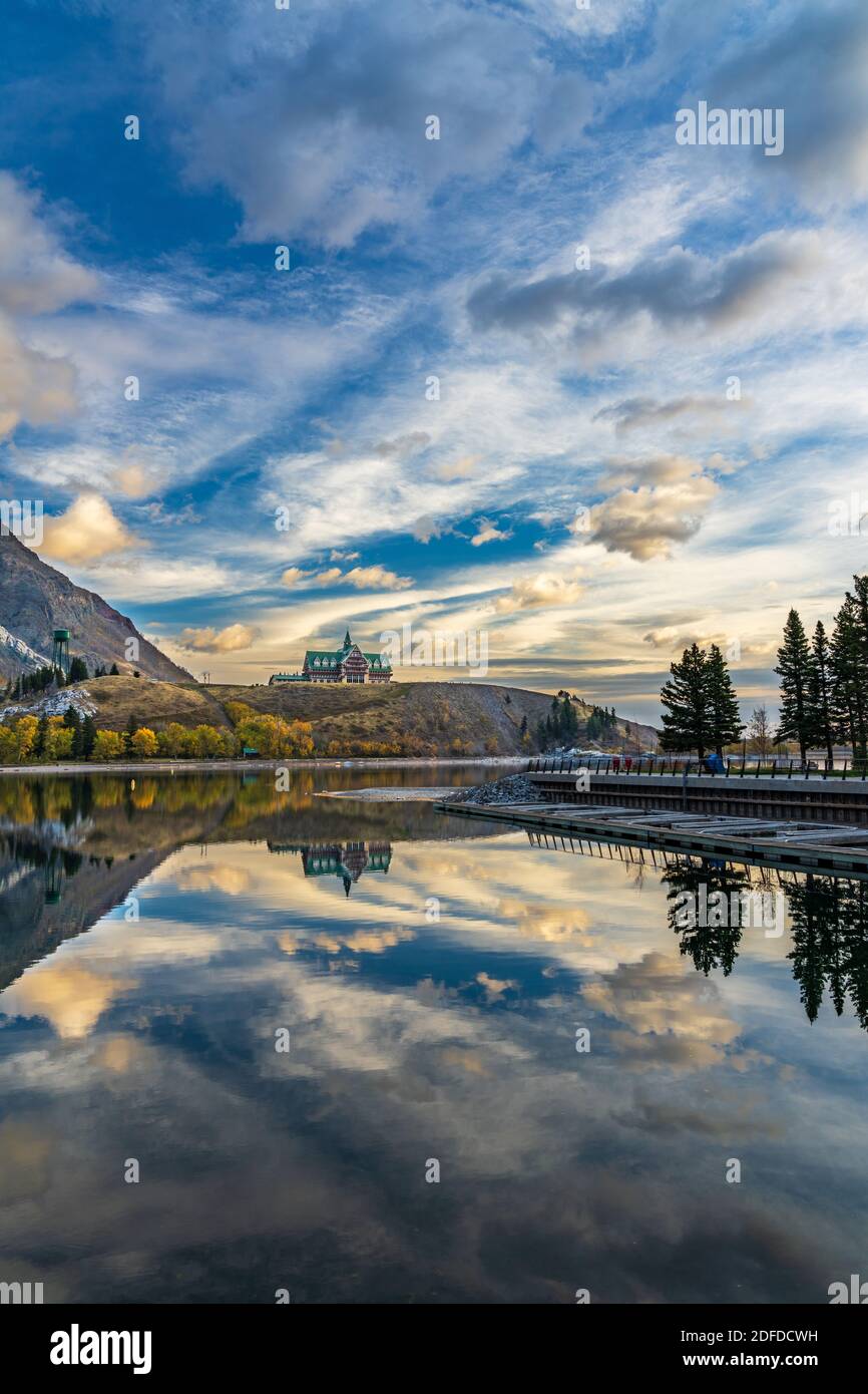 Waterton Lakes National Park lakeshore in autumn foliage season morning. Blue sky, colourful clouds reflect on lake surface like a mirror in sunrise. Stock Photo