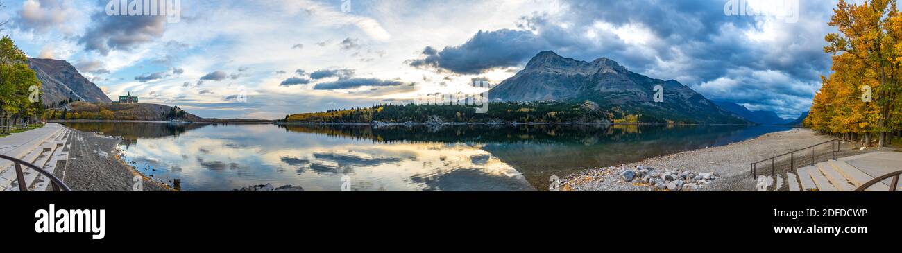 Waterton Lakes National Park lakeshore in autumn foliage season morning. Blue sky, colourful clouds reflect on lake surface like a mirror in sunrise. Stock Photo