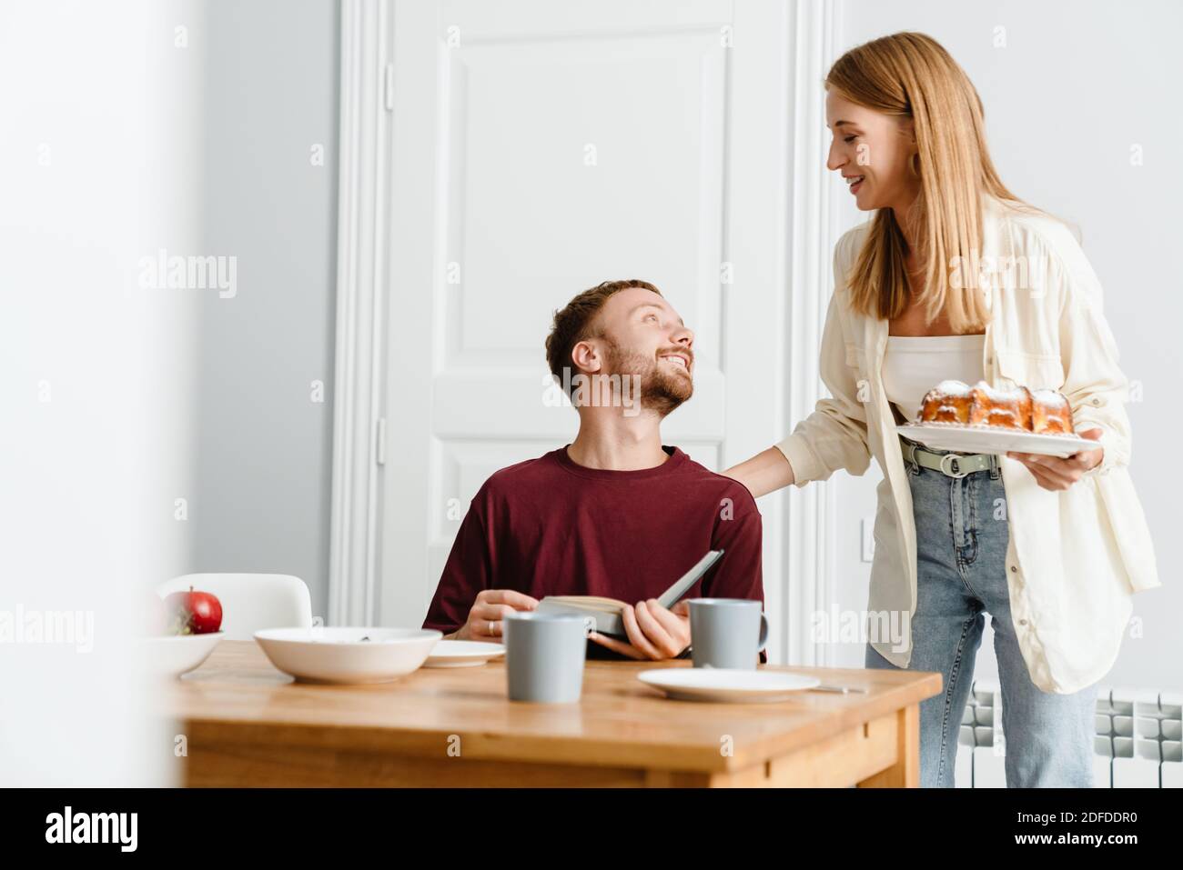 Image of ginger joyful couple drinking coffee with pie while reading book at table in home Stock Photo