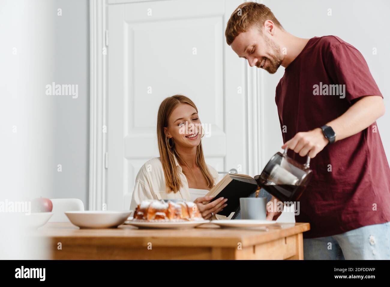 Image of ginger joyful couple drinking coffee with pie while reading book at table in home Stock Photo