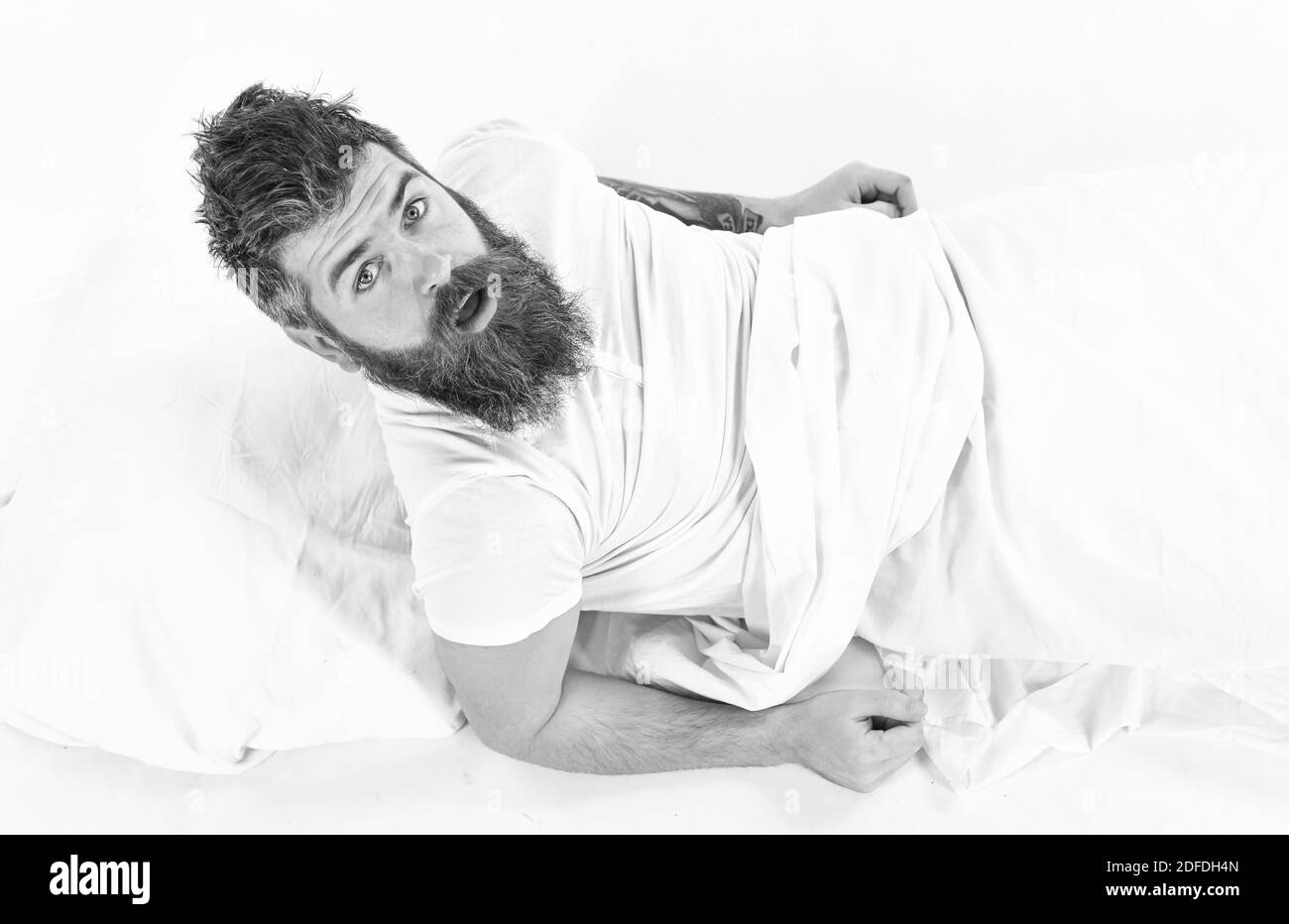 Hipster with beard and mustache waking up. Morning and wake up concept. Man on surprised face, morning, need to get up, white background. Man with sleepy face sit in bed, white sheets, top view. Stock Photo