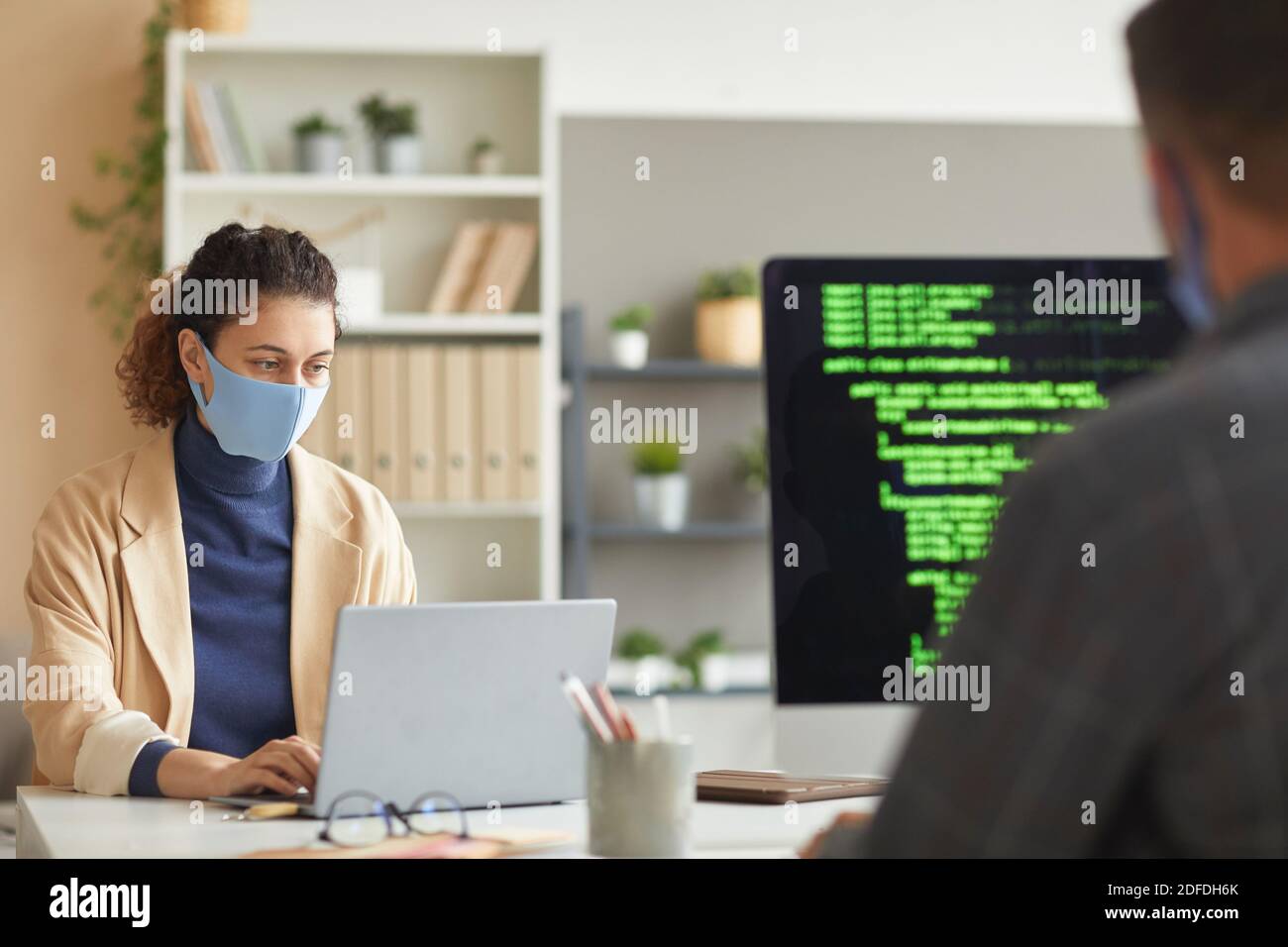 Developers in masks working at the table on computers with codes at office Stock Photo