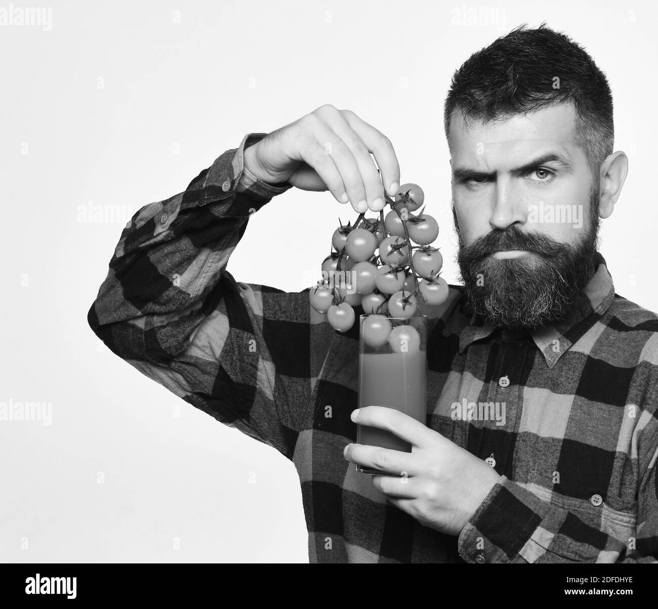 Farmer with serious face shows bunch of red cherry tomatoes. Man with beard holds glass of juice and vegetables isolated on white background. Guy holds homegrown harvest. Farming and autumn concept. Stock Photo