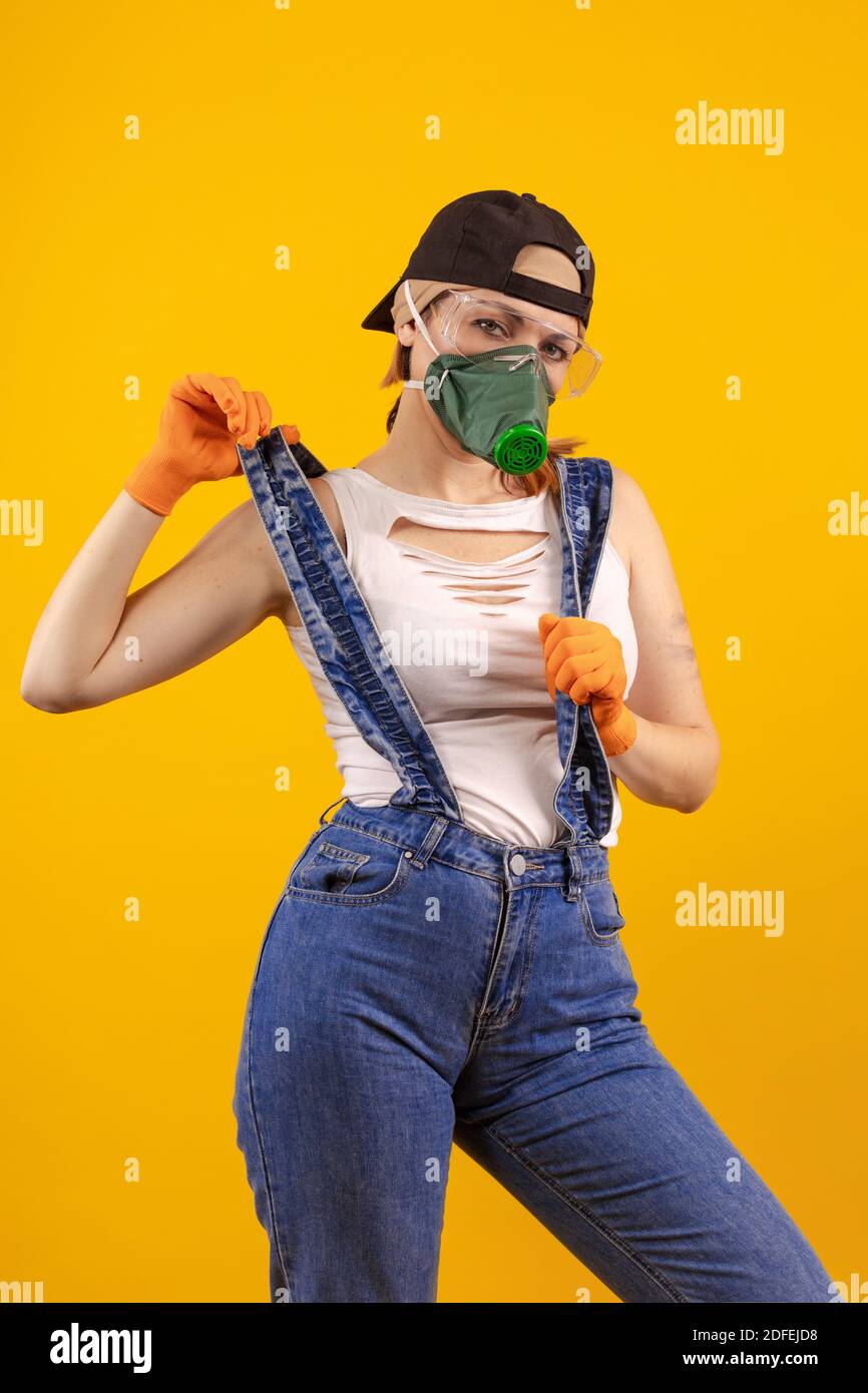 Builder girl in respiratory mask in denim overalls and a black cap posing on a yellow background Stock Photo