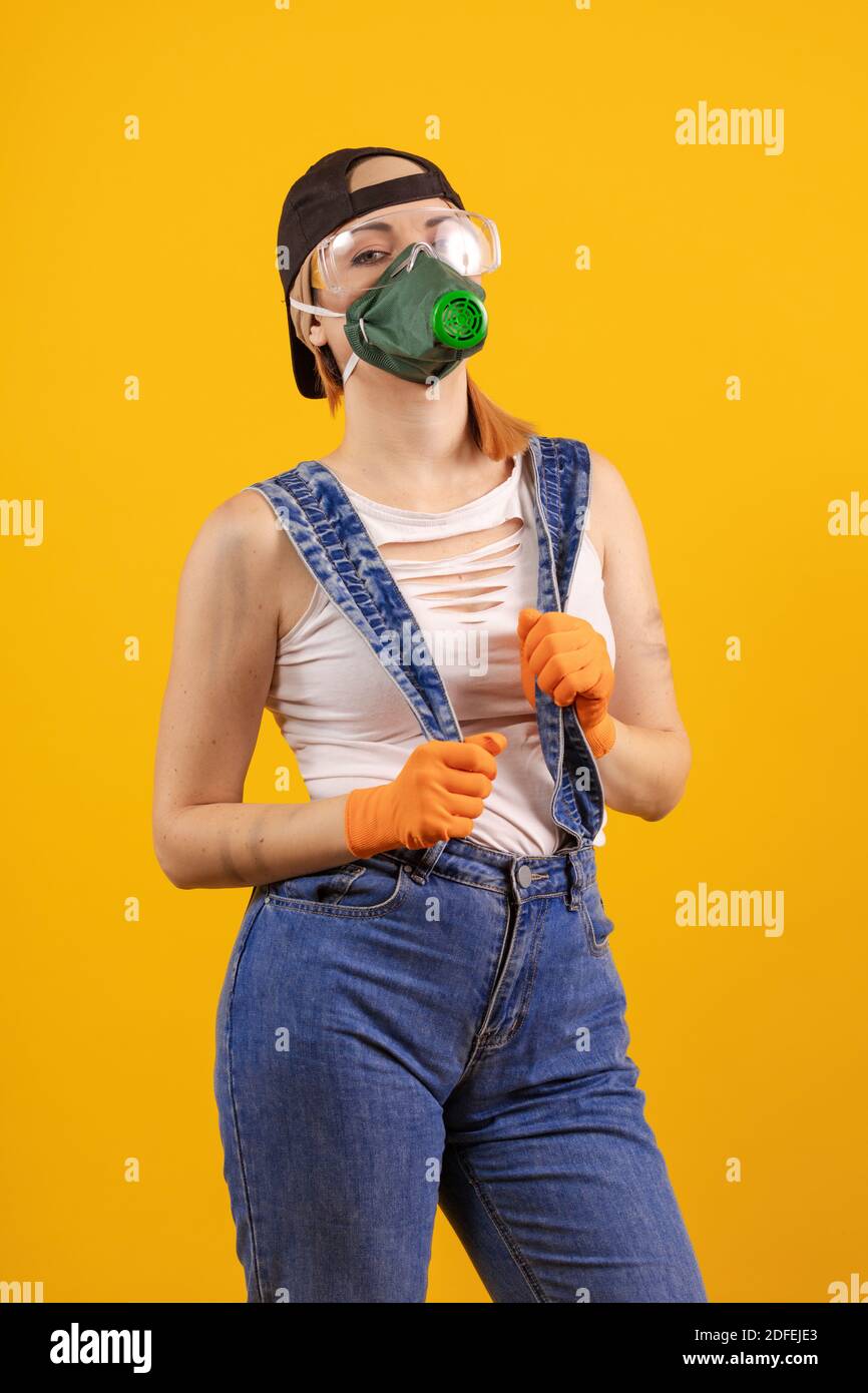 Builder girl in respiratory mask in denim overalls and a black cap posing on a yellow background Stock Photo